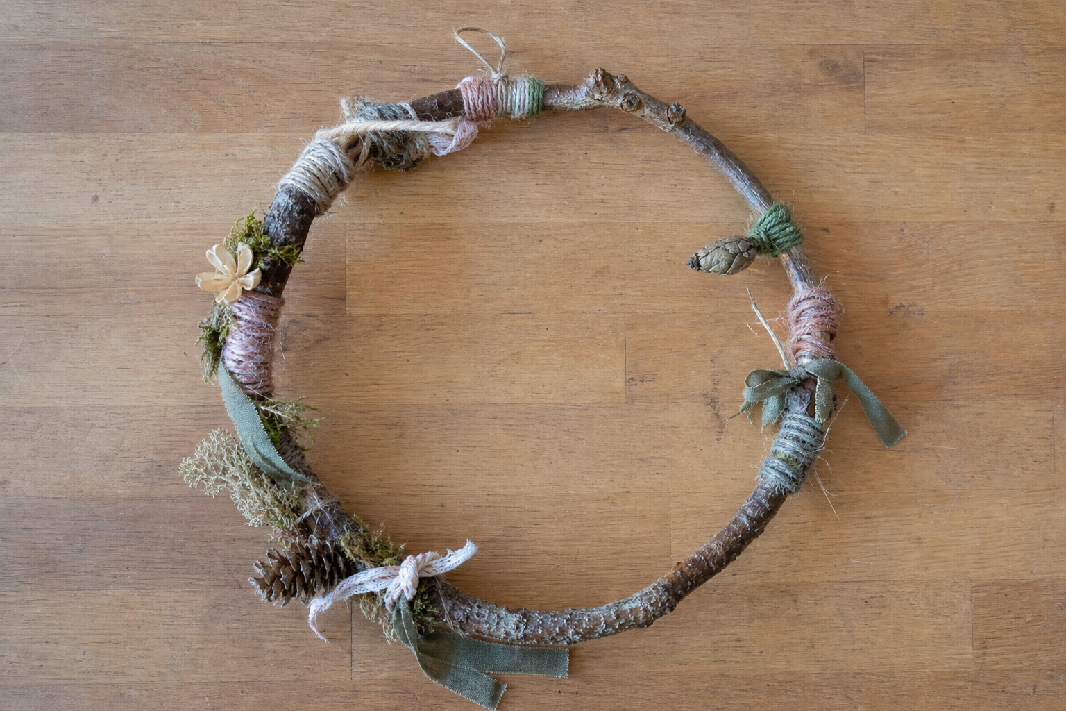 Ein ringförmig gebogener Ast mit Garn, Bändern und dekorativen Naturmaterialien liegt auf einer Holzfläche.